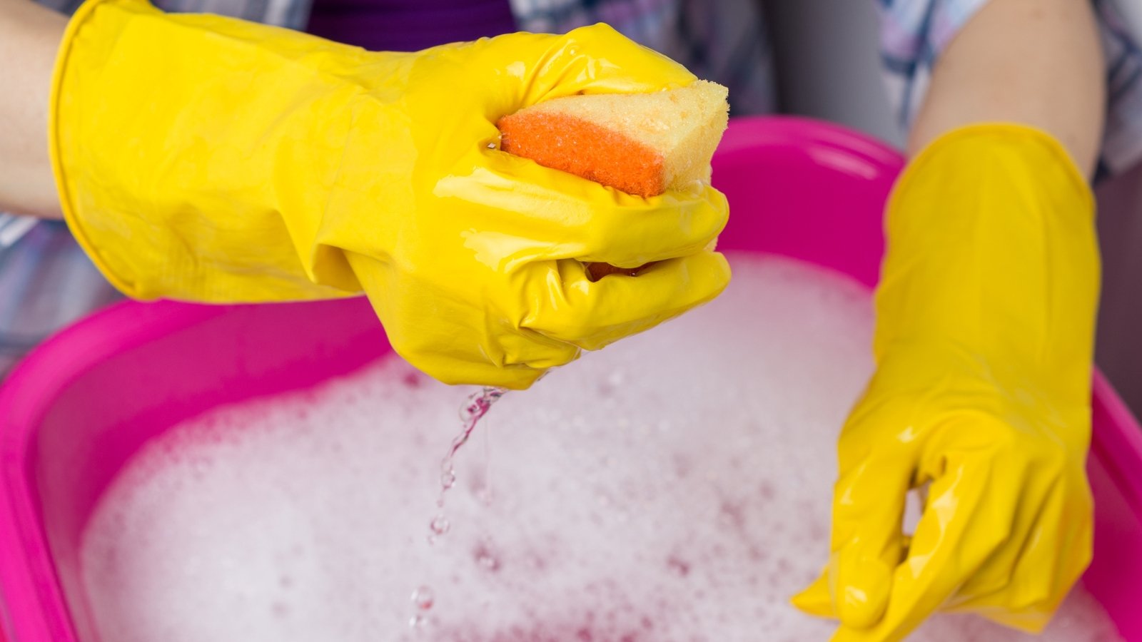 A yellow-gloved hand holding an orange sponge above a pink tub filled with soapy water.